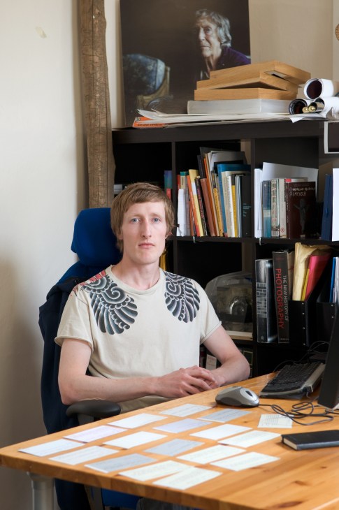 Thomas Østbye sits at his desk at Plymserafin with books and design materials behind him.