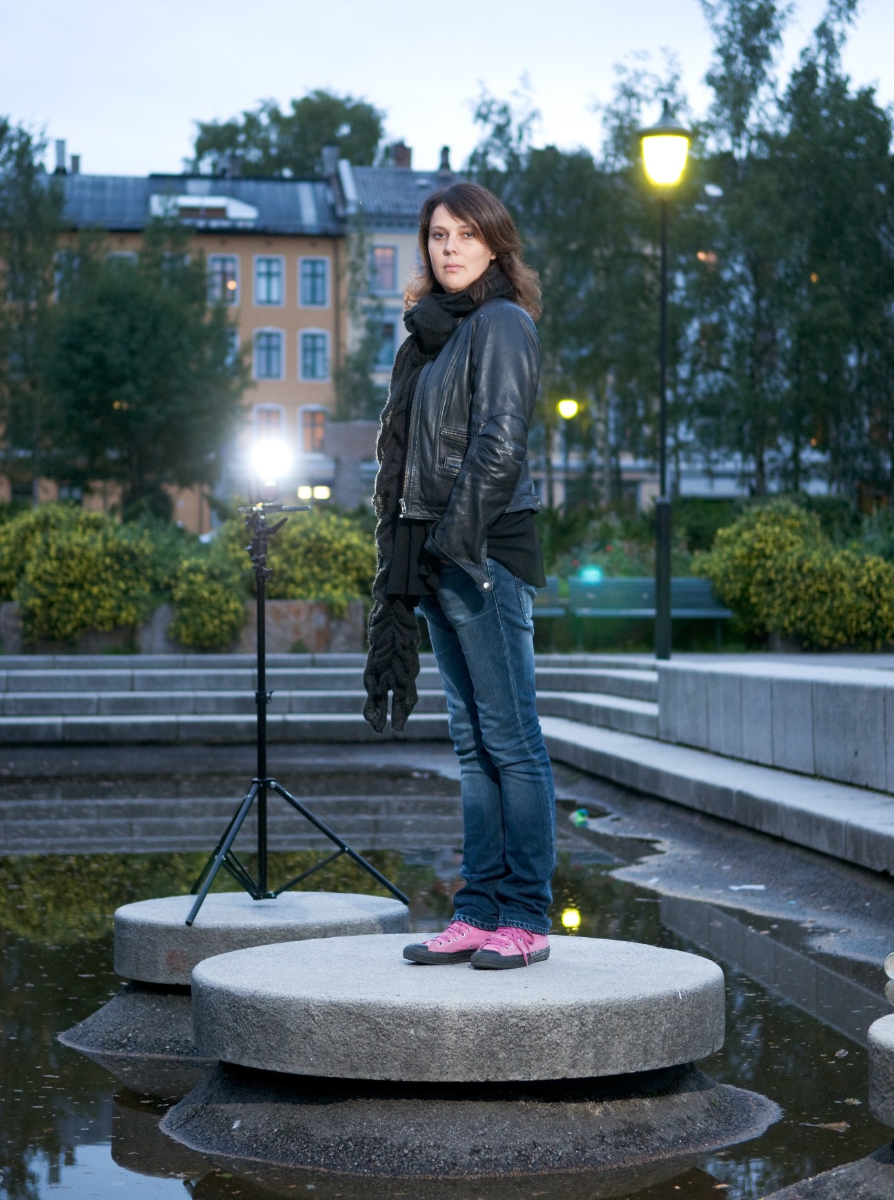 Director Margreth Olin stands on a concrete platform in an Oslo park at dusk, wearing a black leather jacket and pink shoes.