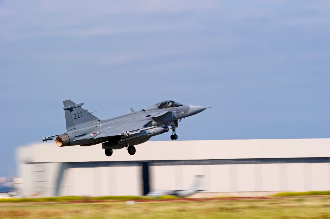Swedish Gripen fighter jet taking off with landing gear down and afterburners engaged.