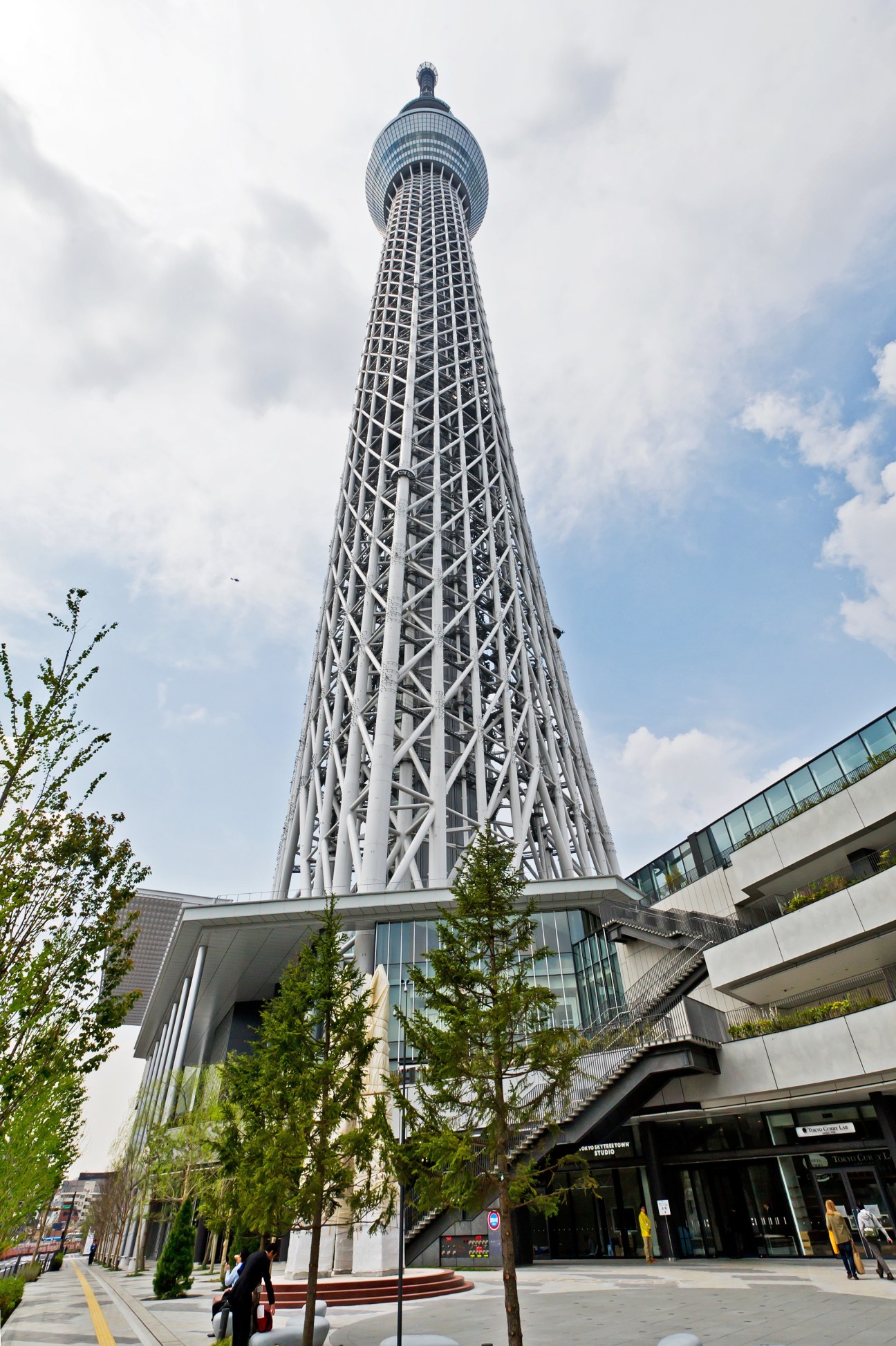 Tokyo Skytree tower with white steel lattice framework rising above modern base building and plaza with trees.