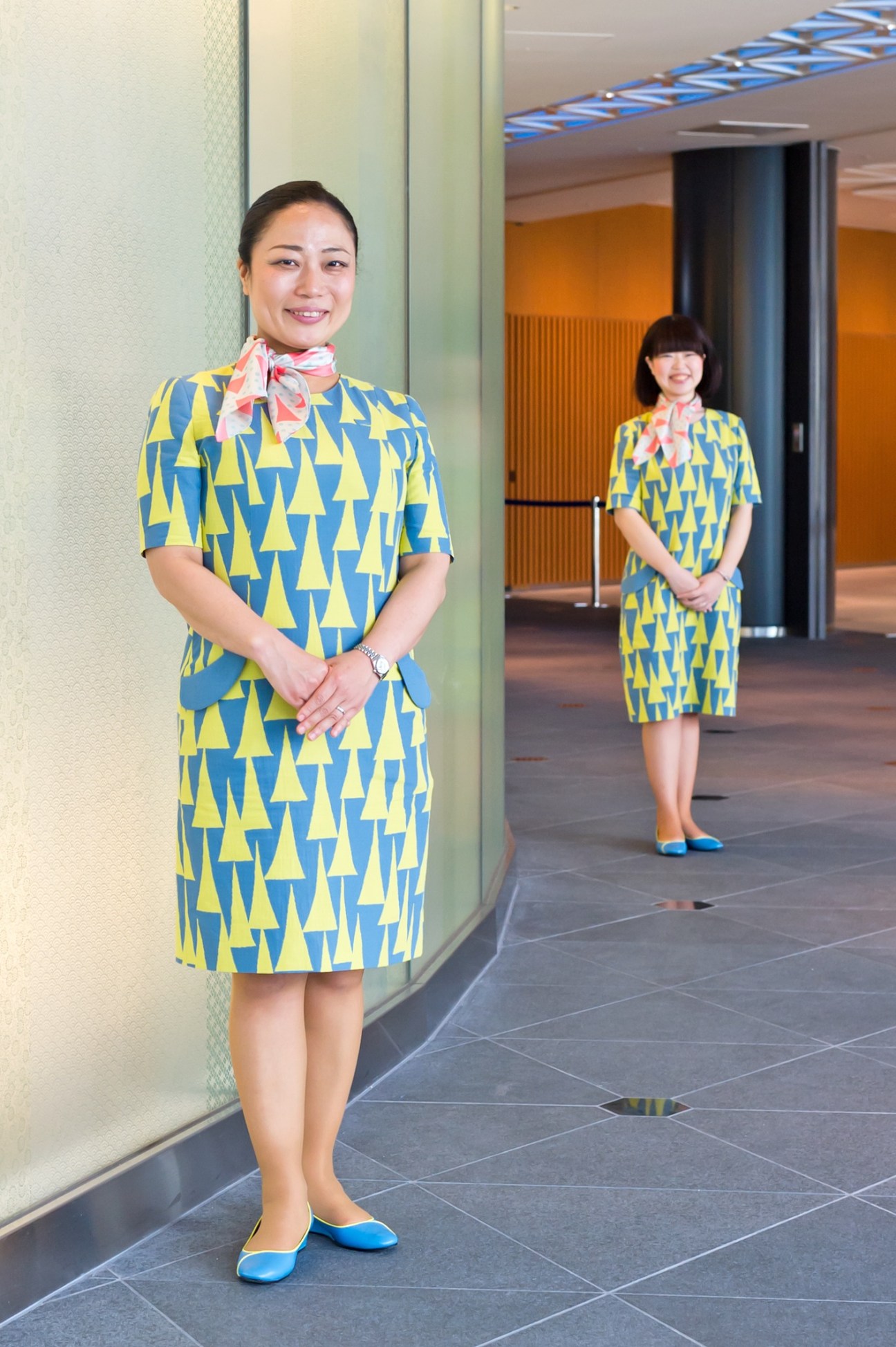 Two female Tokyo Skytree guides wearing blue and yellow patterned dresses designed by Akira Minagawa with colorful scarves and blue shoes.