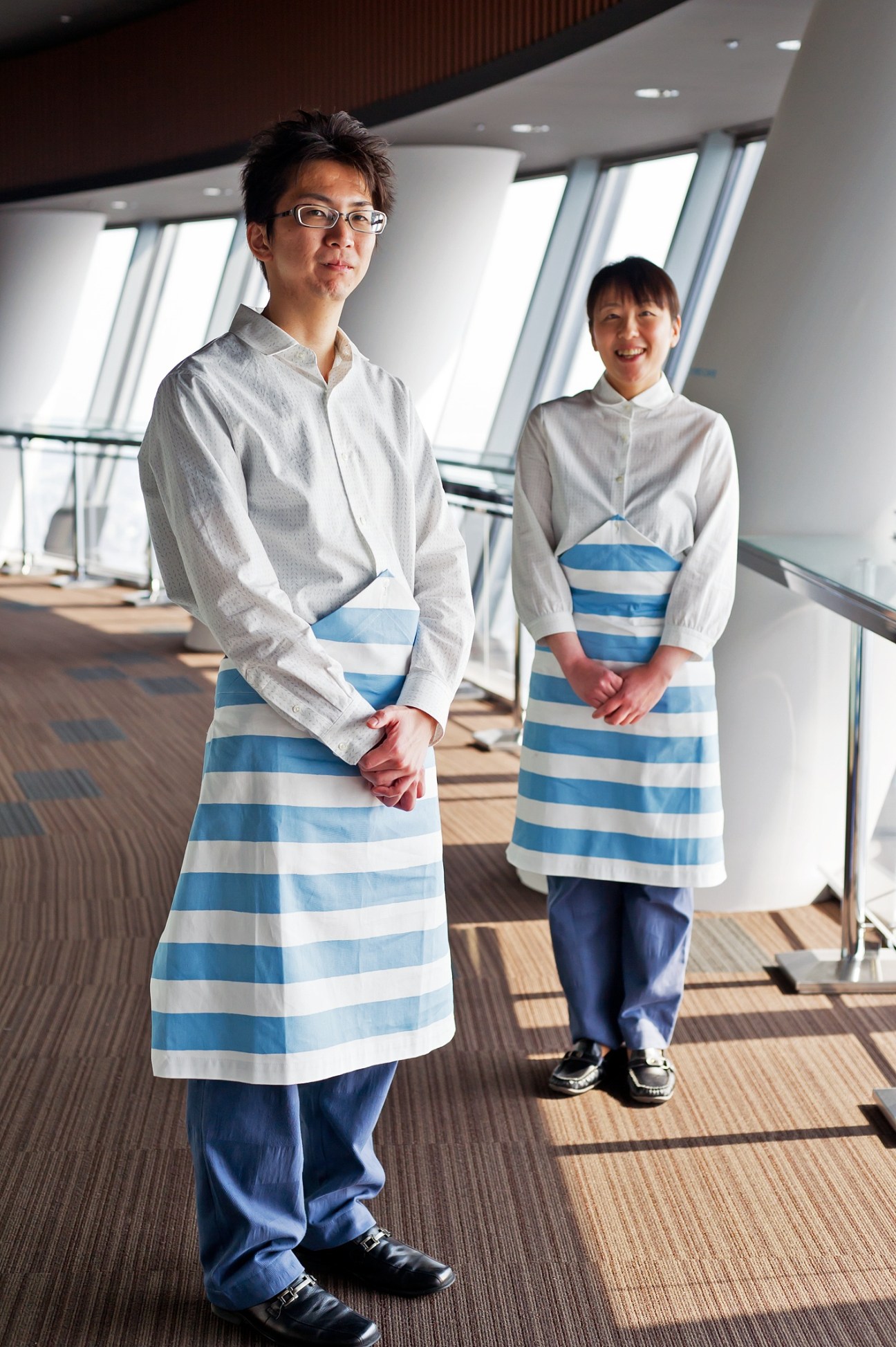Two Tokyo Skytree café staff members wearing blue and white striped aprons over light colored shirts in a modern observation deck interior.