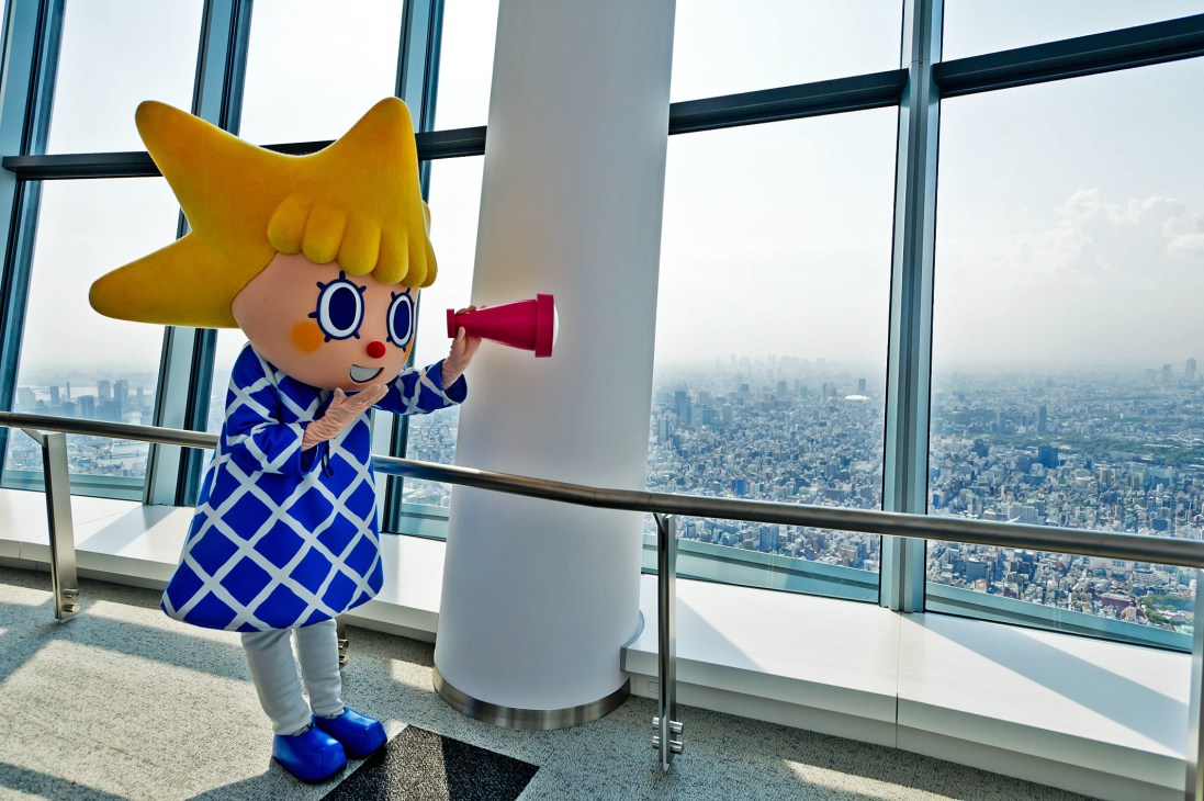 Sorakara, the Tokyo Skytree mascot character, holds a red telescope while standing at an observation deck window overlooking the city.