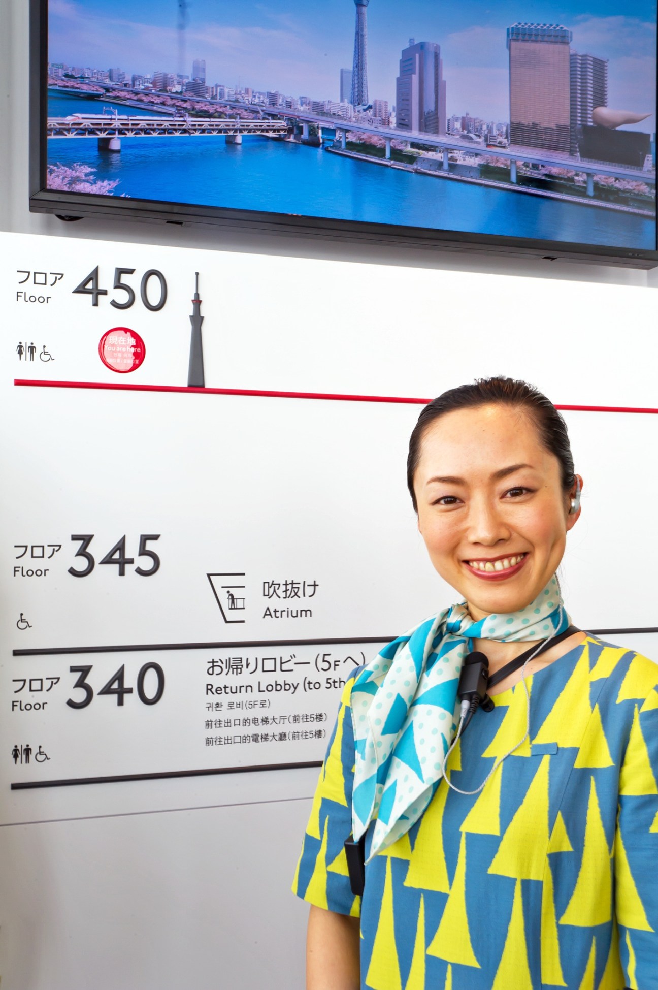 Tokyo Skytree staff member in yellow and blue patterned uniform designed by Akira Minagawa, standing beside navigation signage.