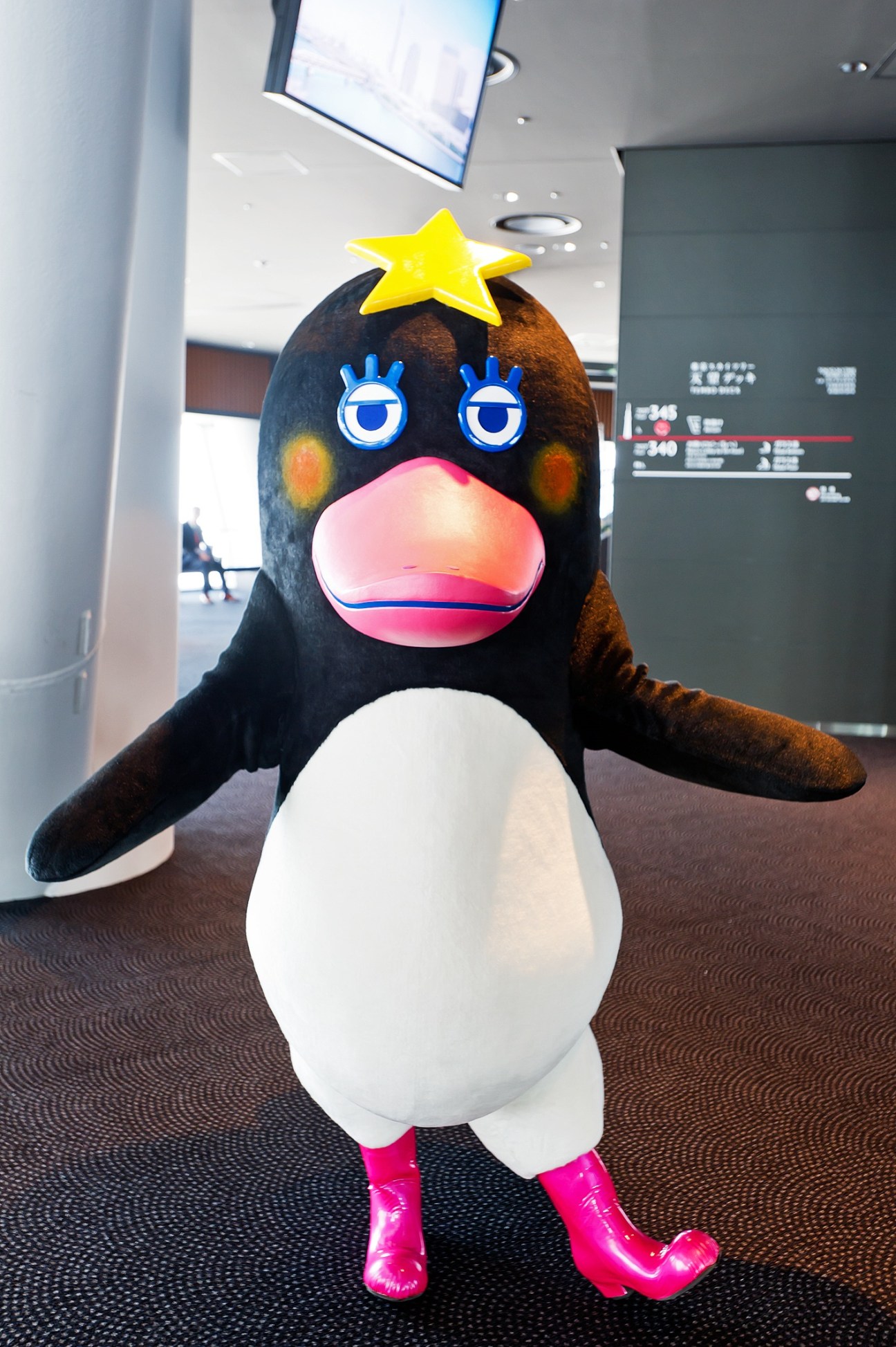 Sorakara mascot character with black body, white belly, pink mouth, blue eyes, yellow star on head, and hot pink feet standing indoors.