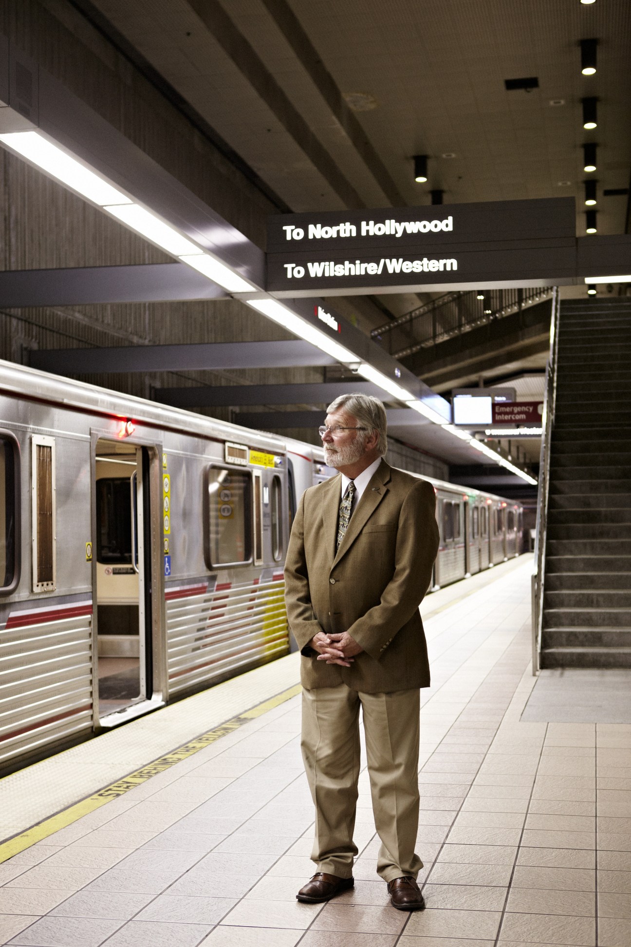 Art Leahy stands on a Los Angeles metro platform wearing a tan suit, with a train and directional signs visible behind him.