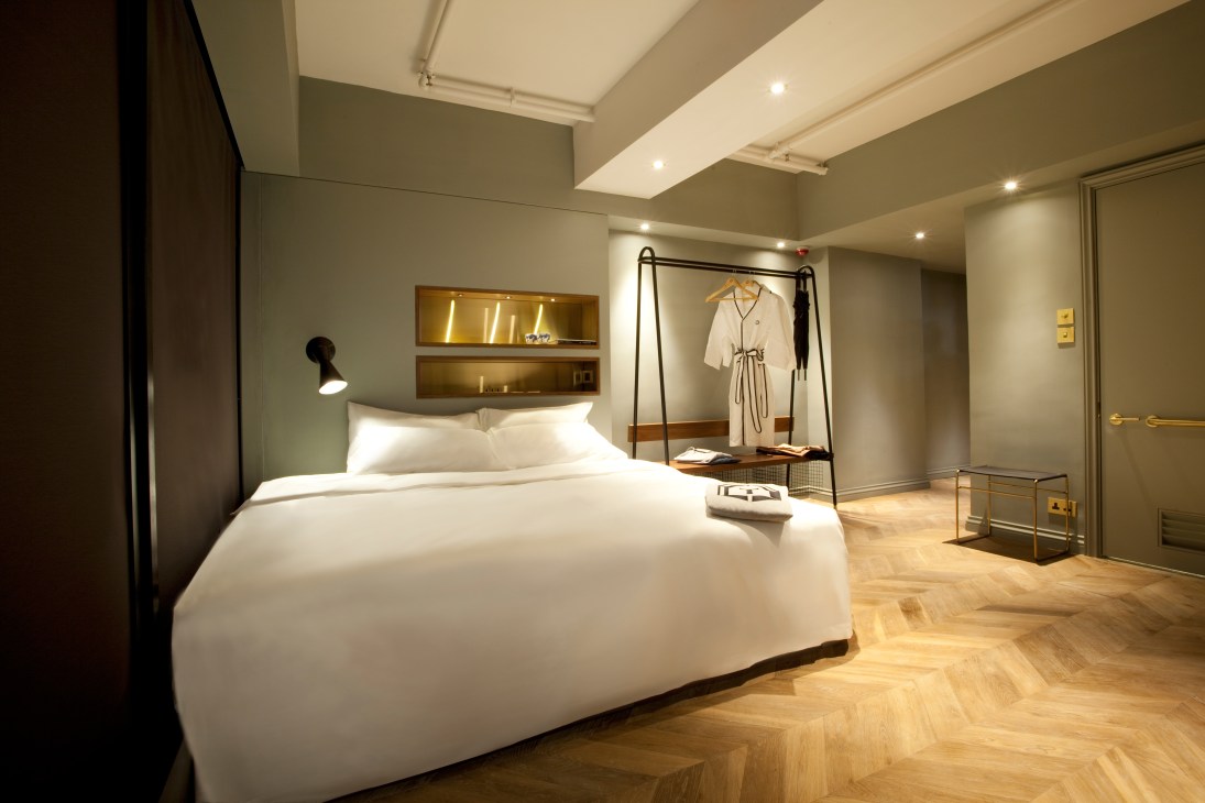 Contemporary hotel bedroom with white bedding, sage green walls, built-in shelving, and wooden herringbone flooring at Tribute.