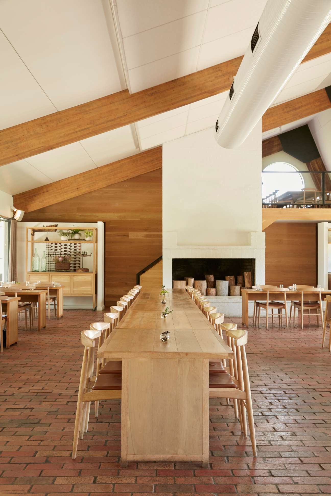 A long communal wooden dining table with light wood chairs in a bright, modern restaurant space with exposed timber beams and brick flooring.