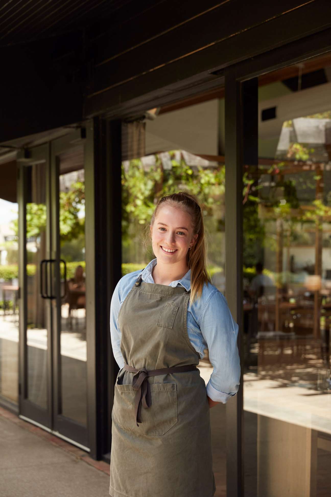 Young female restaurant employee in khaki apron and blue shirt standing in front of Mitchelton's modern glass and timber architecture.