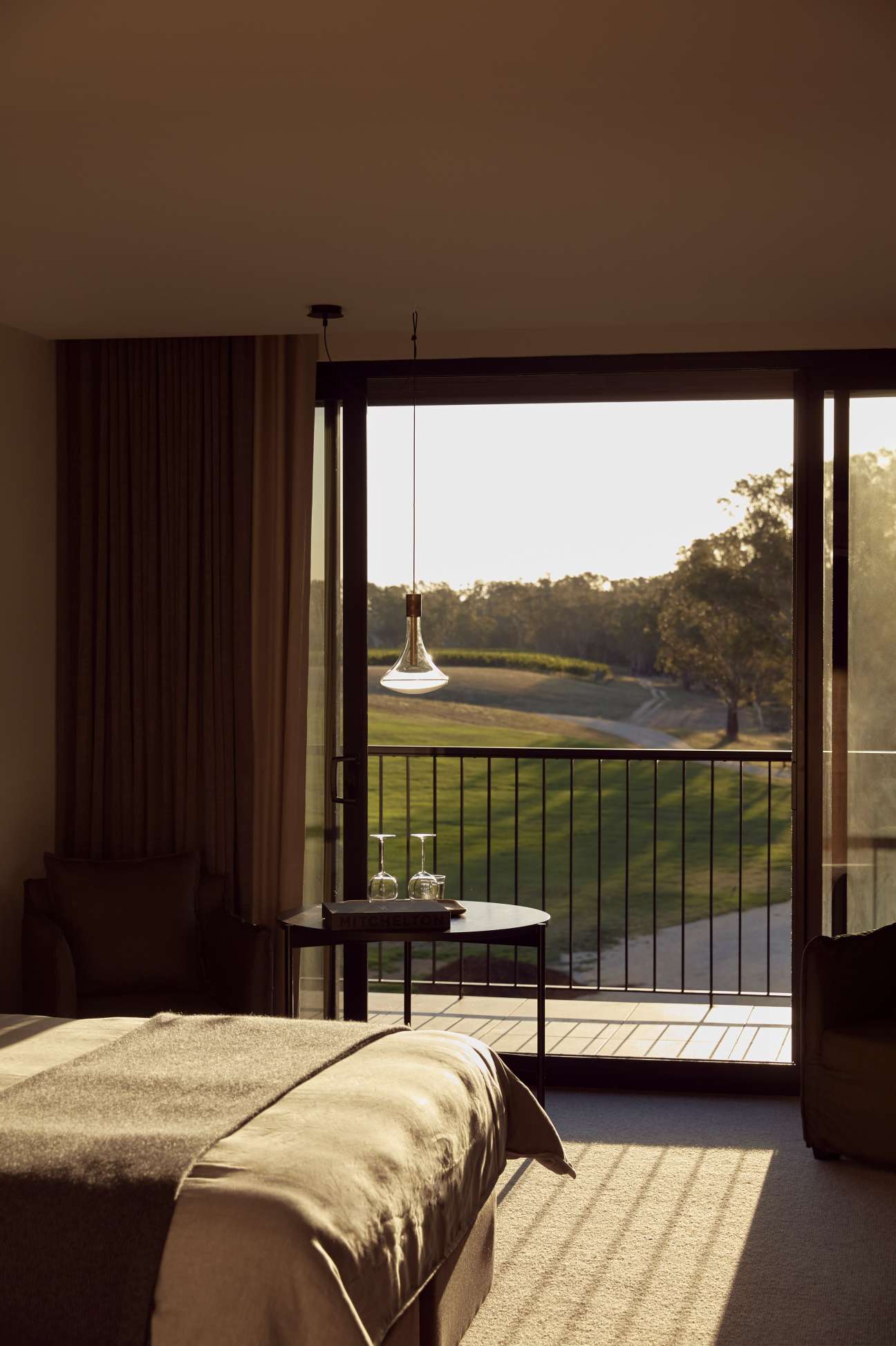 Hotel bedroom with vineyard views through glass doors and a pendant light over a side table.