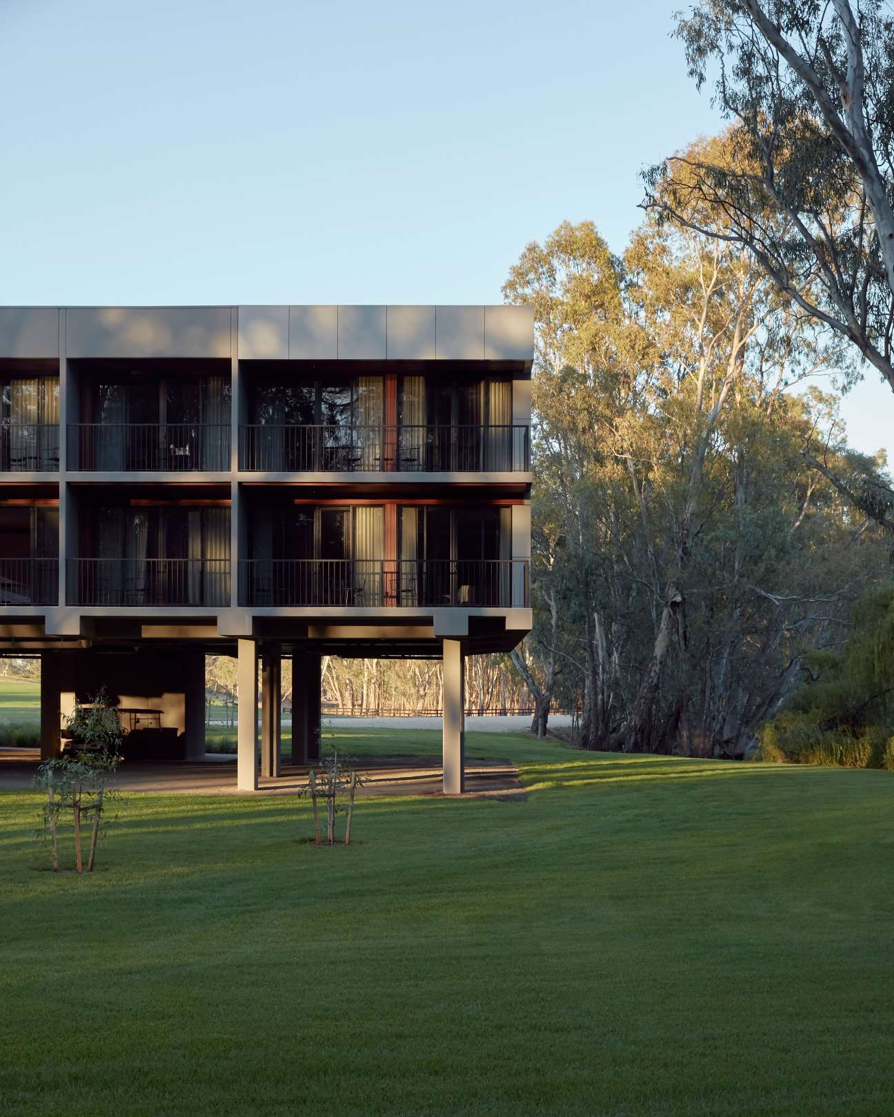 Modern two-story hotel building with horizontal lines and balconies overlooking manicured lawn and river with eucalyptus trees.