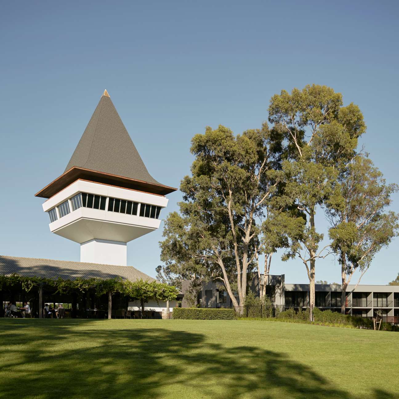 Mitchelton's distinctive lookout tower with conical roof stands in a grassy courtyard beside gum trees and mid-century buildings.