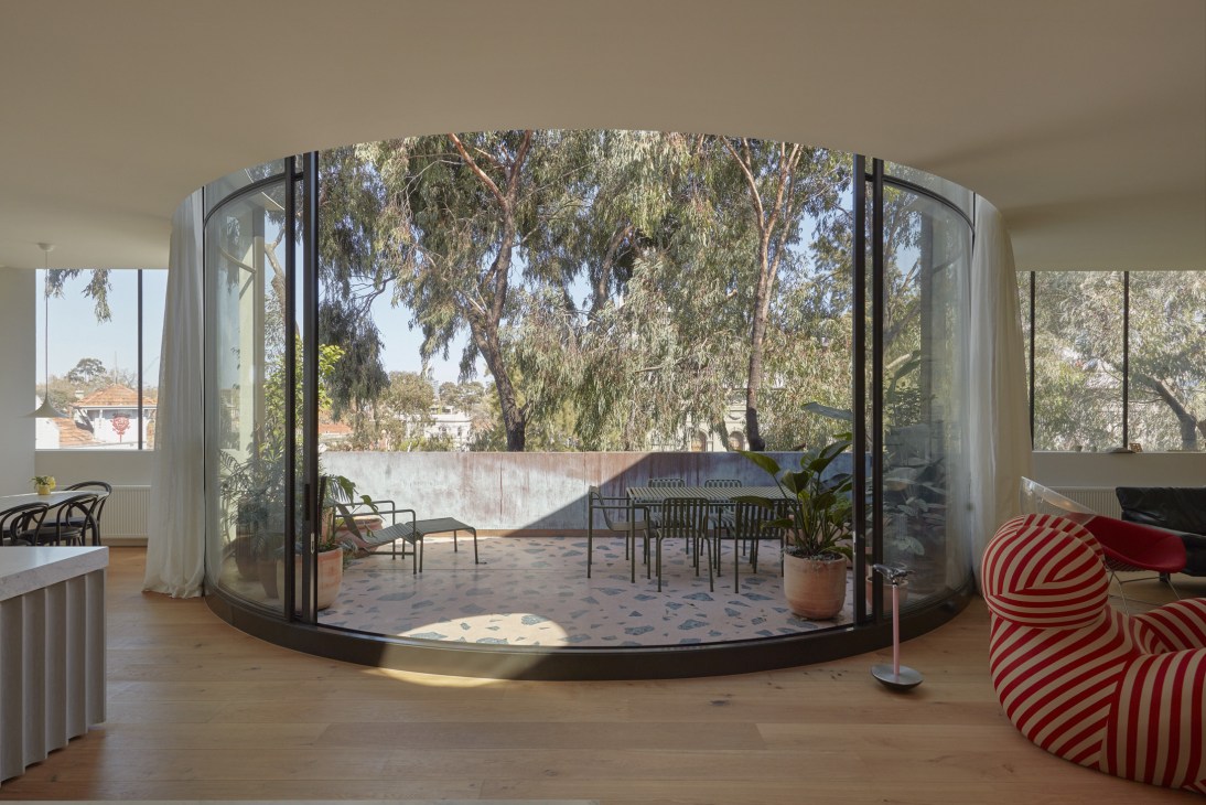 Curved glass doors opening onto a terrace with eucalyptus trees and city views beyond, featuring minimalist outdoor furniture.