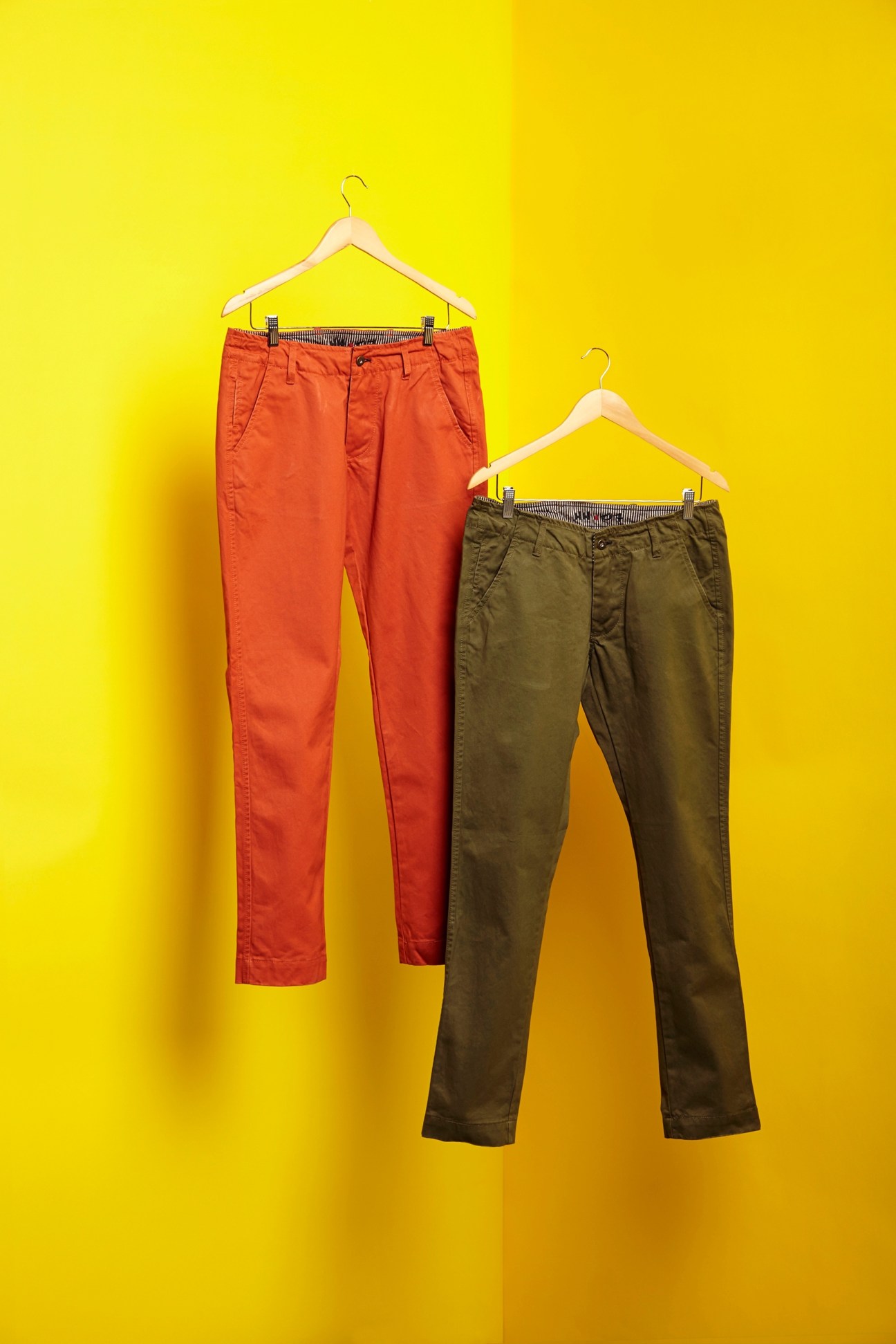 Two pairs of NN.07 chinos displayed on wooden hangers against a yellow background—one in coral red and one in olive green.
