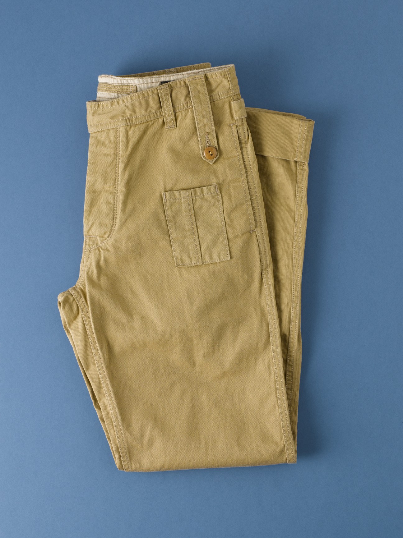 Folded beige utility trousers with cargo pockets against a blue background.