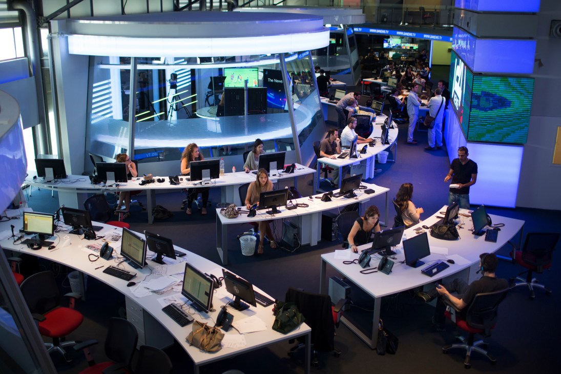 Overhead view of i24 News editorial floor with journalists working at curved desks surrounded by multiple monitors and screens.