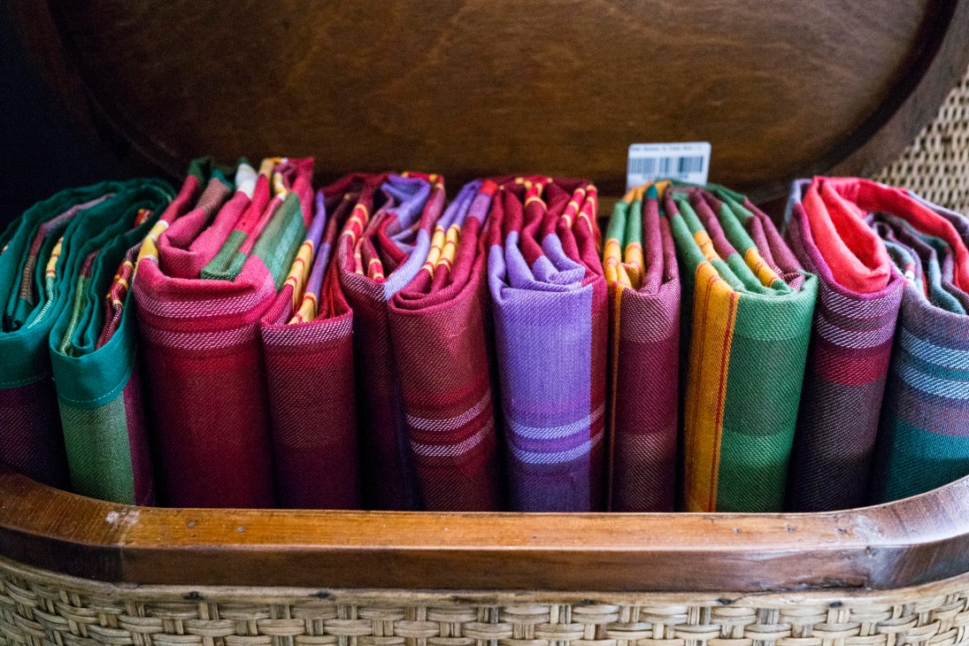 Colorful rolled textiles and fabrics in vibrant pink, purple, green and gold tones displayed in a wooden basket.