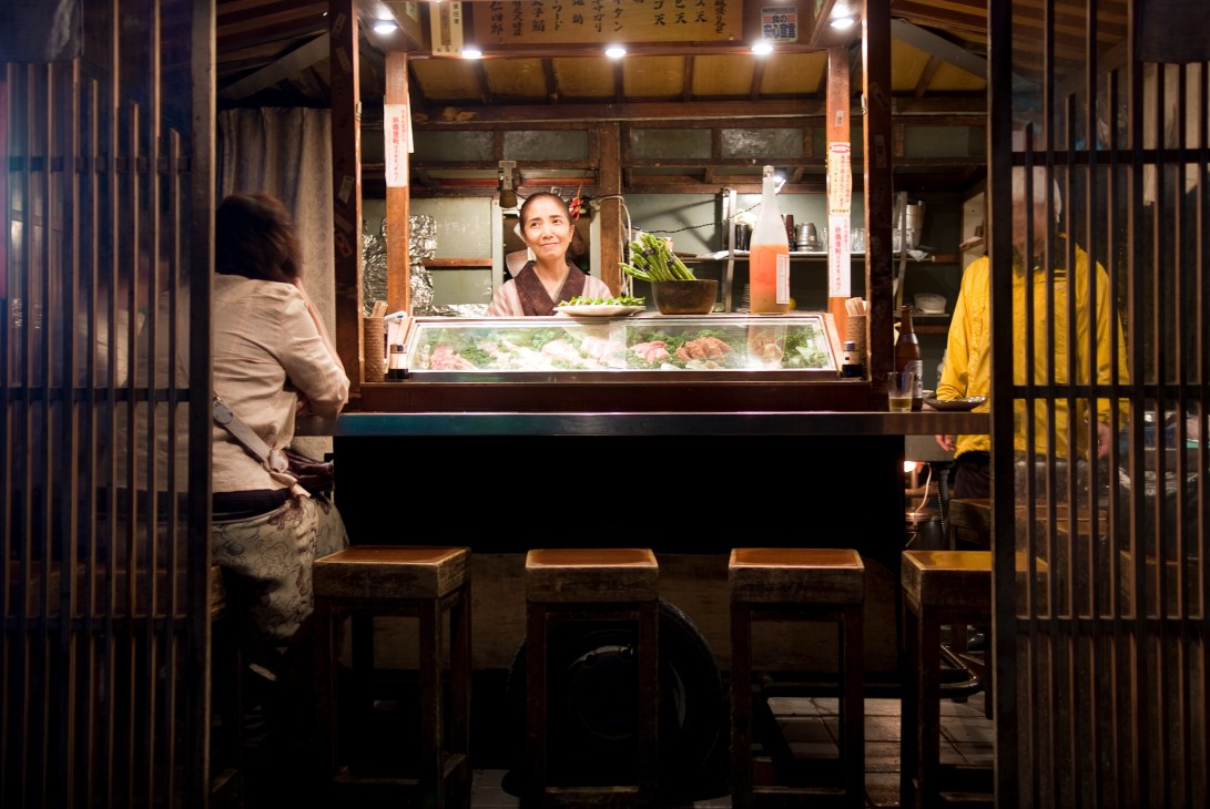 A vendor serves food from a traditional yatai food stall with wooden counter and stools.