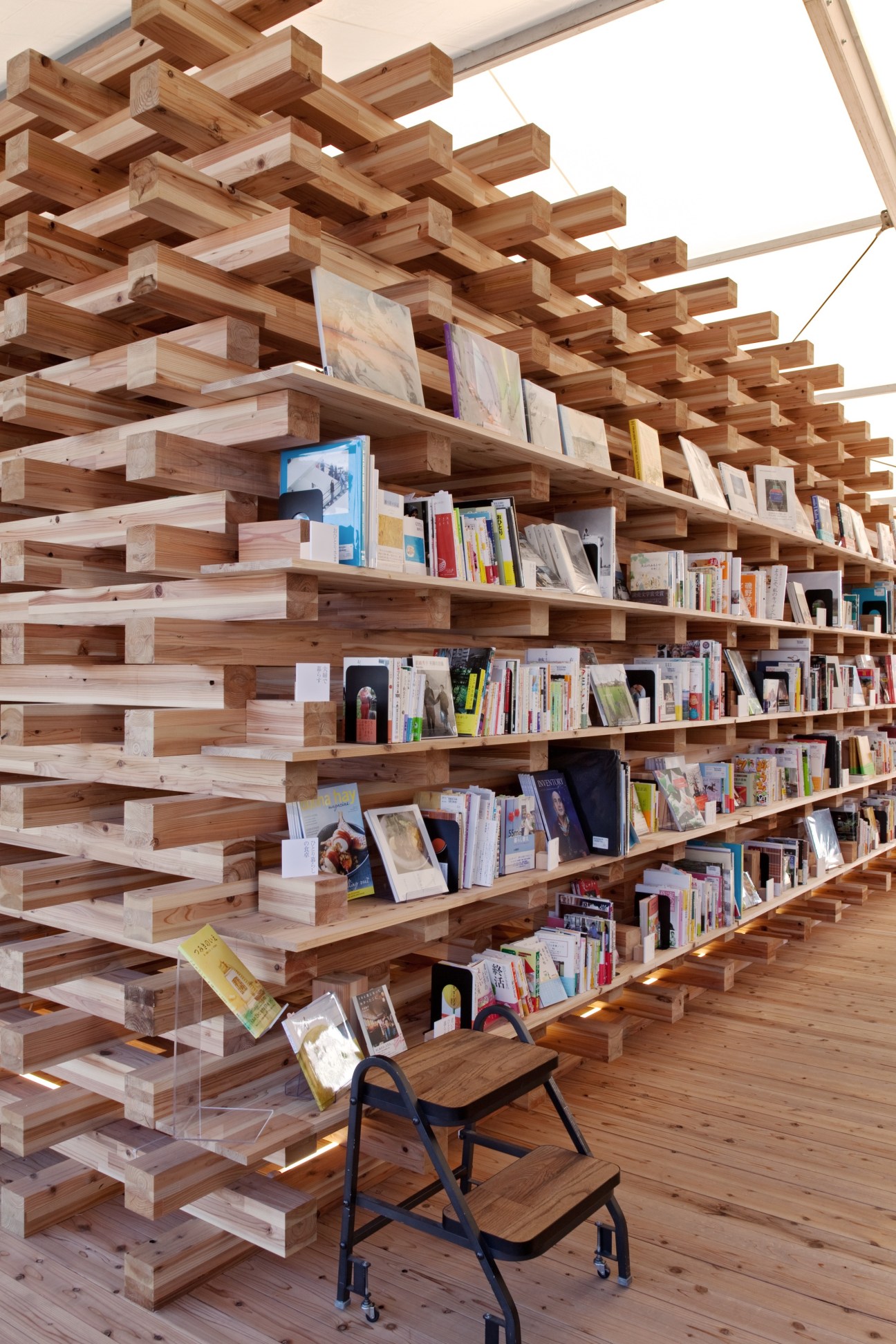 Wooden shelving system with stacked beams displaying books and magazines, with a blue and black chair in foreground.