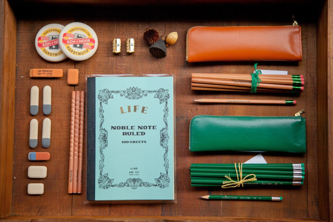 Flat lay of stationery items including notebooks, pencils, erasers, leather wallets and brushes on wooden surface.