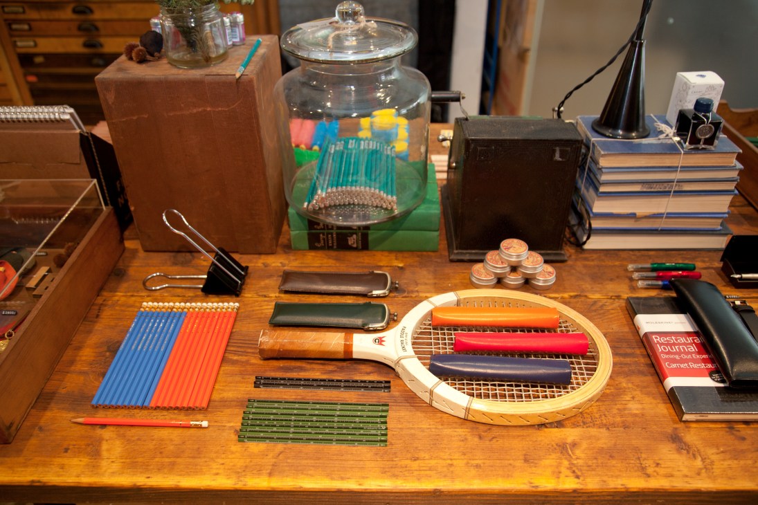 Wooden desk display at Tools to Live By shop showing colorful pencils, leather wallets, tennis racket, stationery and desk accessories.