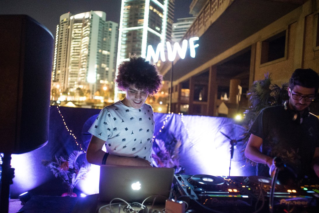DJ performing at night on a rooftop terrace with illuminated purple lighting and city buildings visible in the background.