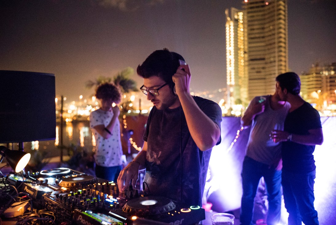 DJ operating turntables at a nighttime rooftop party overlooking illuminated city buildings and bay.