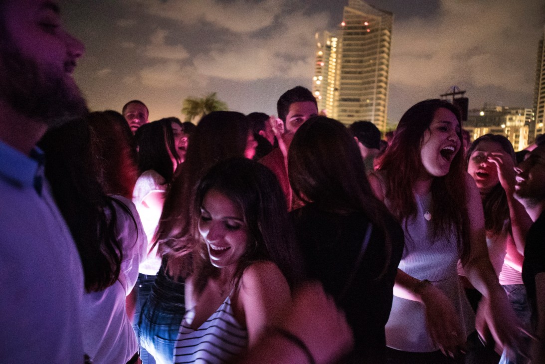Group of people dancing and celebrating on a crowded dance floor at night with city buildings lit up in the background.