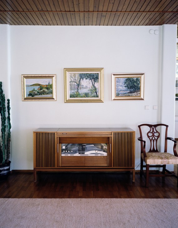 Mid-century wooden stereo console with framed landscape paintings hung above it on a white wall.