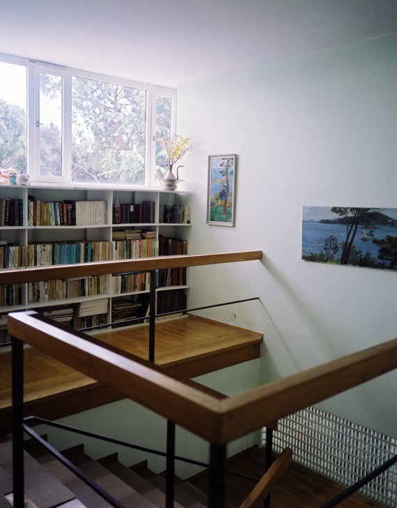Upstairs hallway with wooden staircase, built-in bookshelves, and large windows overlooking pine trees.