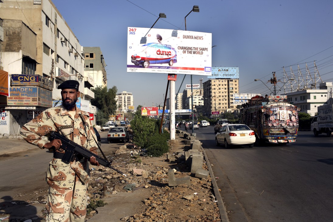 Pakistani ranger in camouflage uniform stands with rifle beneath a Dunya News billboard on a Karachi street.