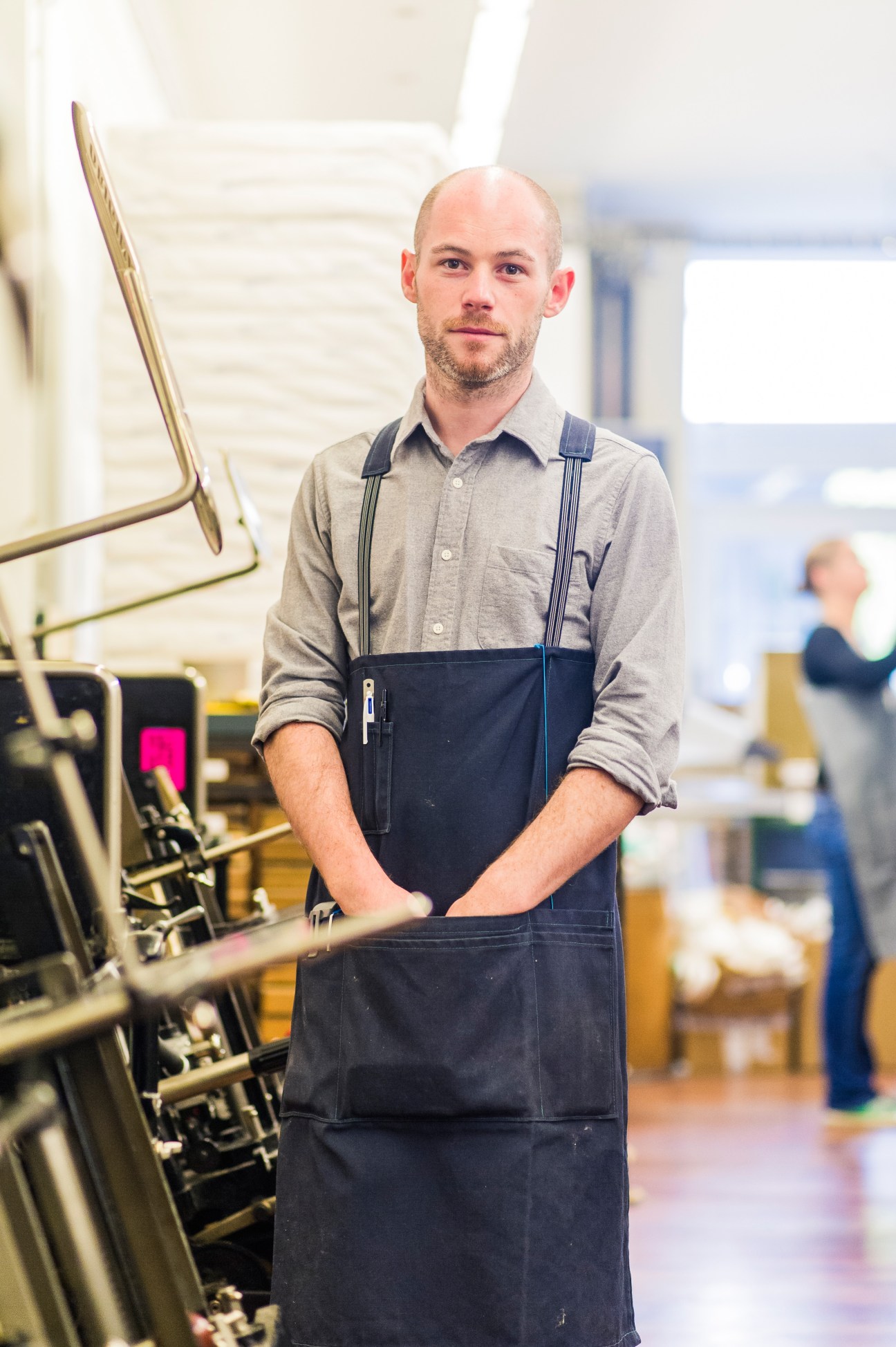 Cédric Chauvelot, founder of Le Typographe, stands in his Brussels printing workshop wearing a gray shirt and dark apron.