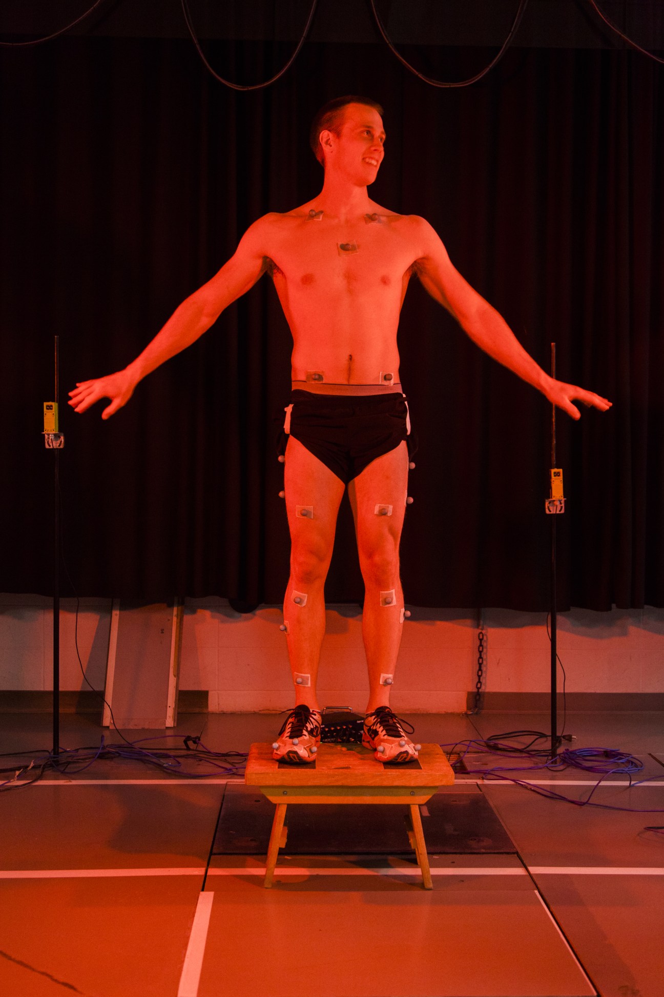 Shirtless male subject with sensors attached standing on stage under red lighting for biomechanics testing.