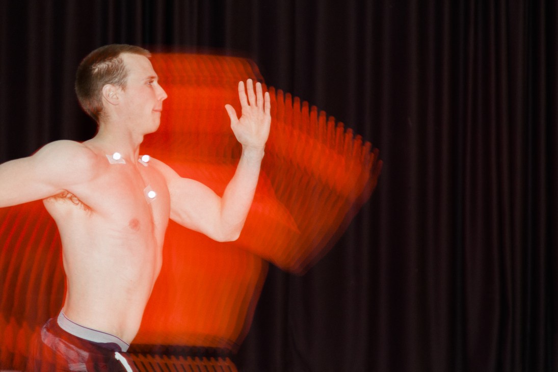 Shirtless male athlete with motion-capture sensors on chest performing running test against red backdrop in laboratory.