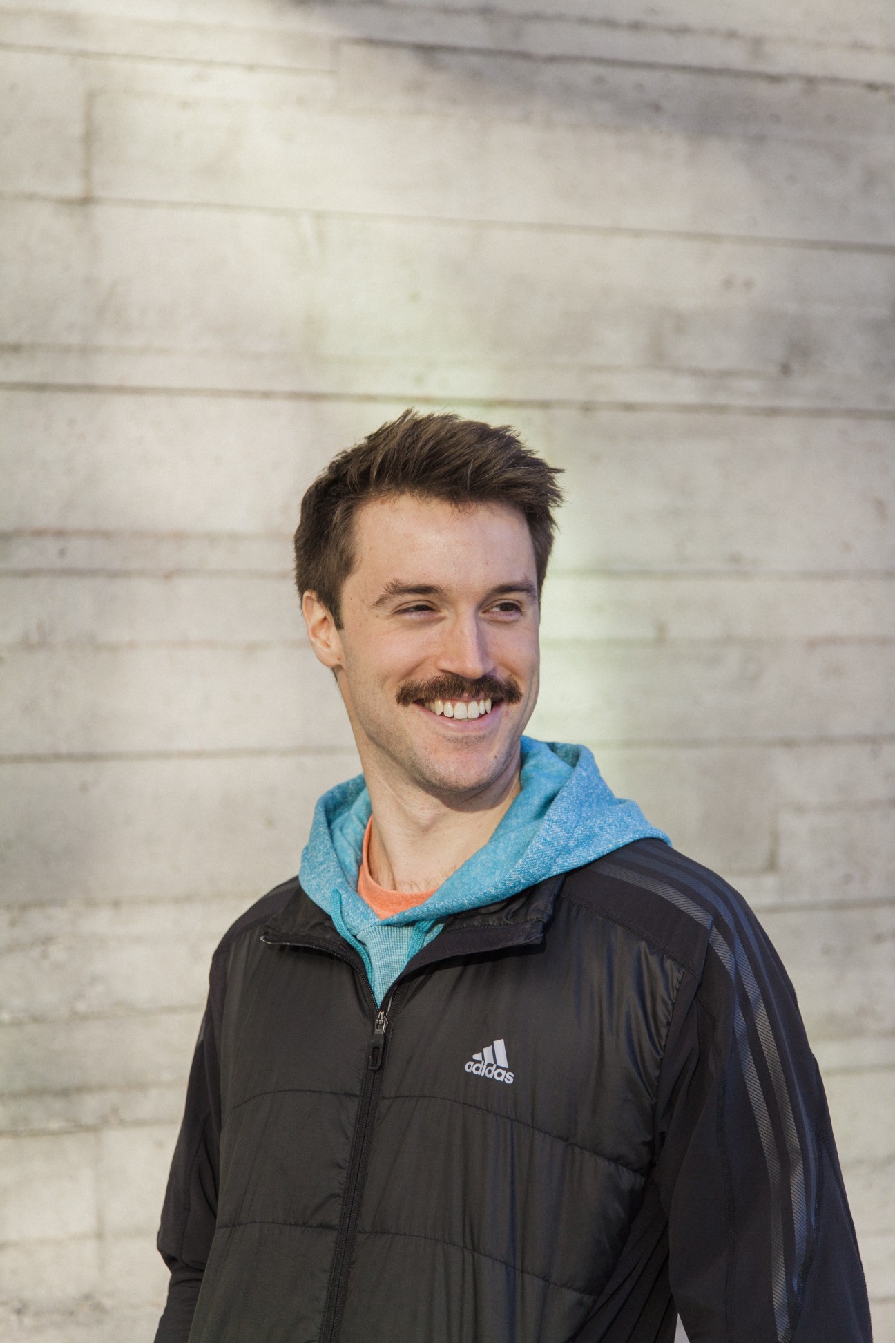 Research assistant Ryan Madden smiling in front of a white brick wall, wearing an Adidas jacket and blue hoodie.
