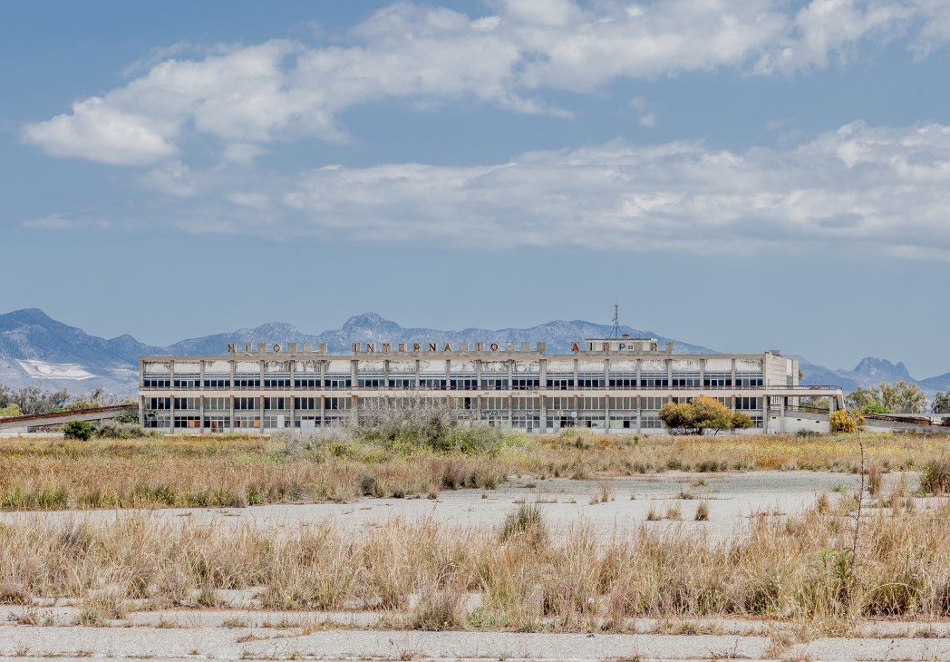 Abandoned Nicosia International Airport terminal building with faded signage surrounded by overgrown vegetation and mountains in background.