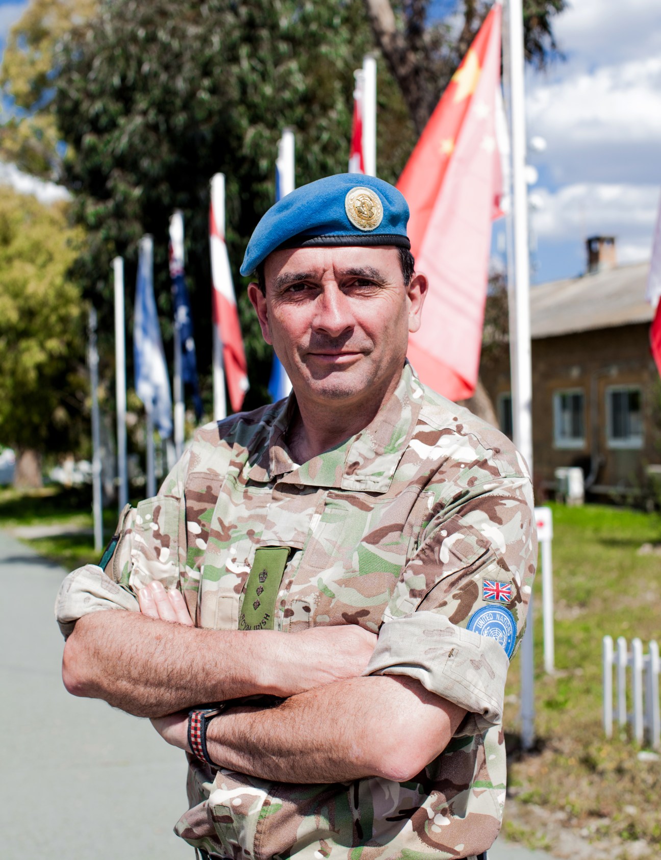 Colonel Angus Loudon of the Royal Irish Regiment in desert camouflage uniform and blue UN beret, arms crossed, standing in front of international flags.