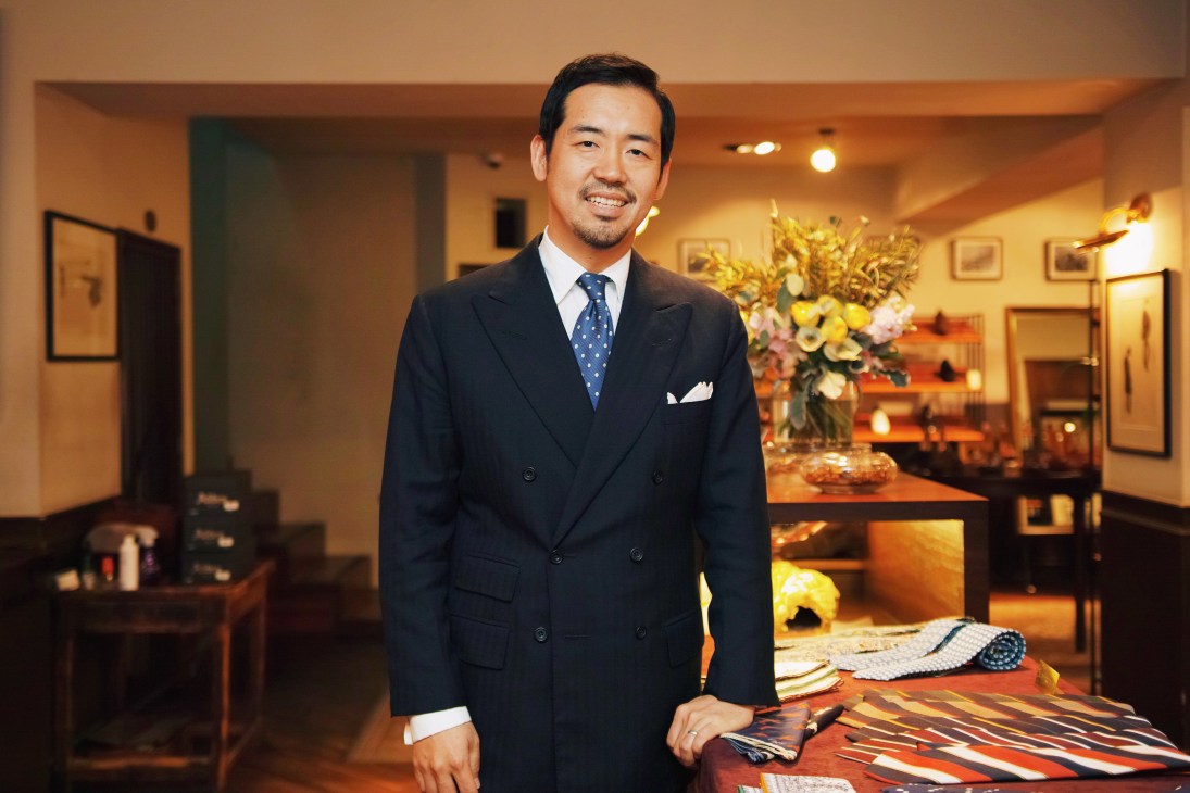 Unipair CEO Jaeyoung Kang stands in a double-breasted navy suit and polka-dot tie in an upscale shoe shop interior.