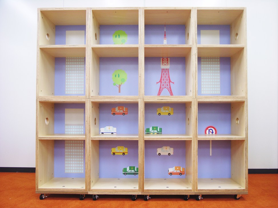 Wooden shelving unit with sixteen compartments displaying colorful illustrated icons of cars, trees, buildings and landmarks on purple backgrounds.