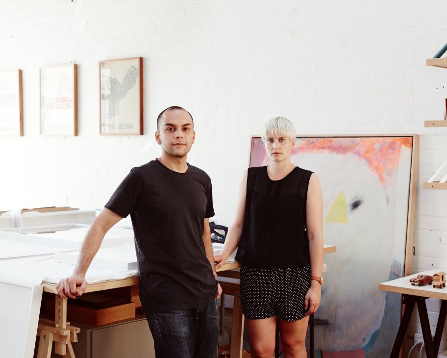 Two designers posing in a bright studio surrounded by wooden picture frames and artwork.