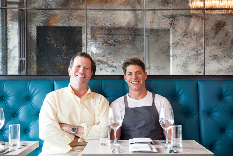Jeffrey Costello and Nick Honeyman sitting at a restaurant table with a blue leather banquette behind them.