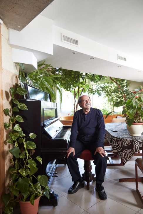 Architect Antonio Cortés Ferrando sits at a piano in his lush apartment surrounded by potted plants and greenery.