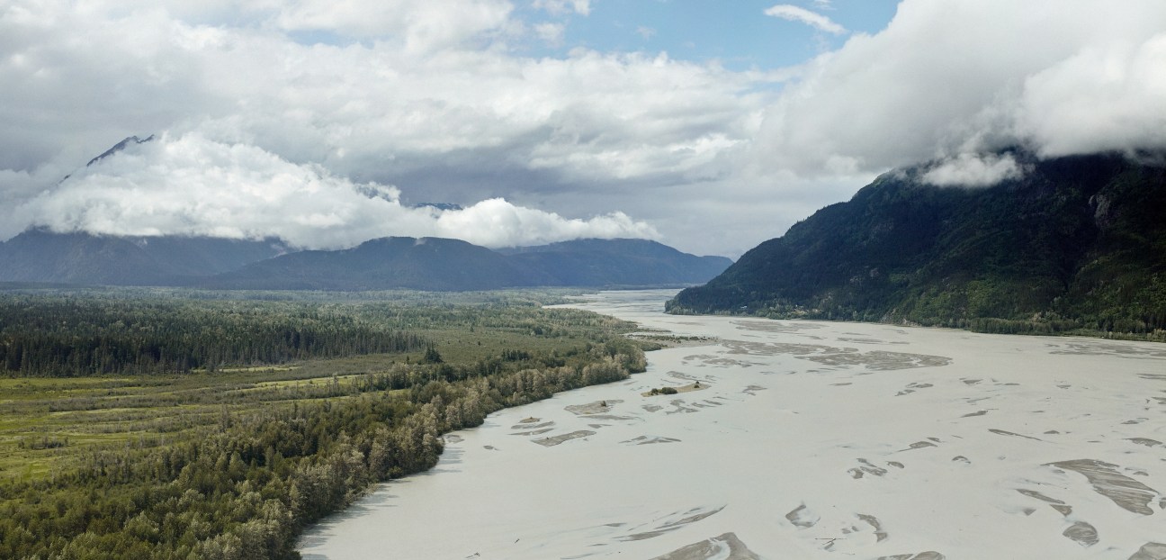 Aerial view of the Chilkat River tidal flats with forested shores, mountains, and cloudy sky.