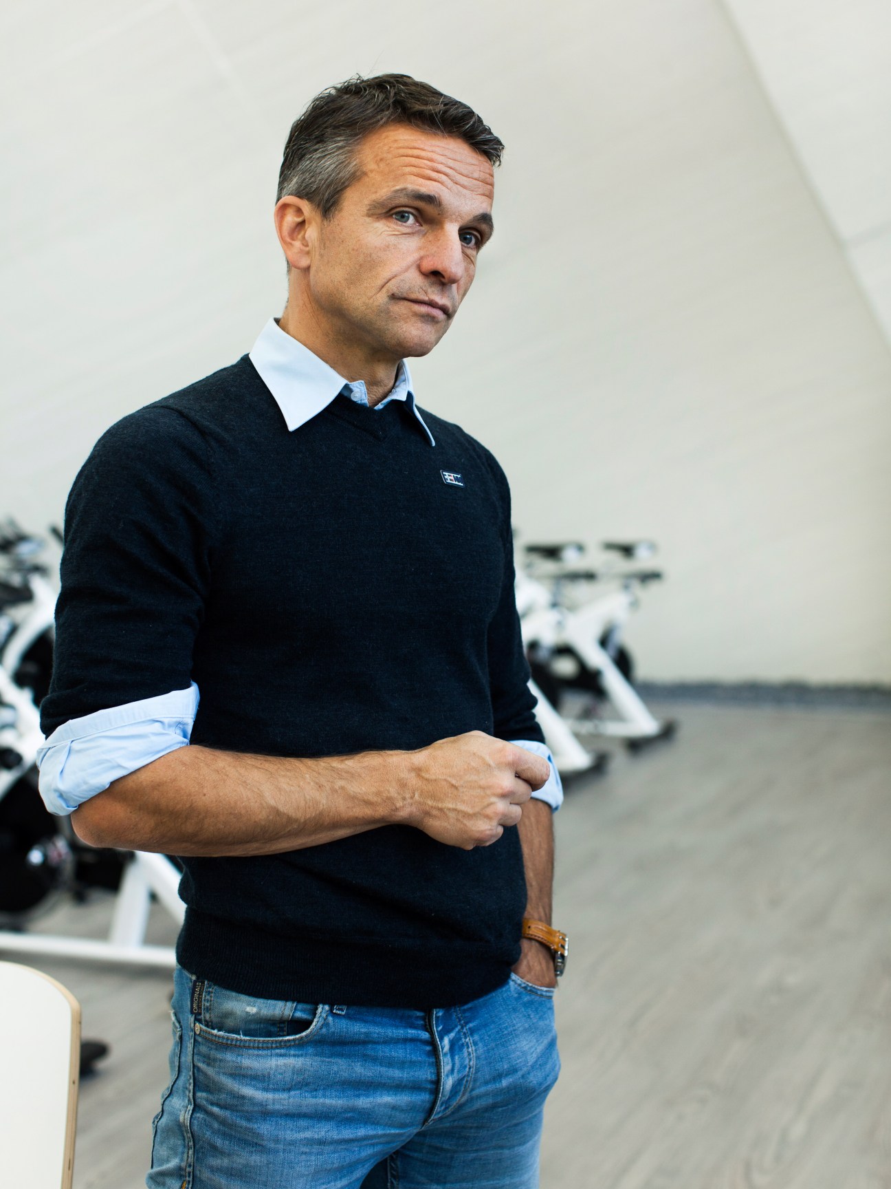 Man in dark sweater and light blue shirt standing in a fitness facility with motorcycles visible in the background.
