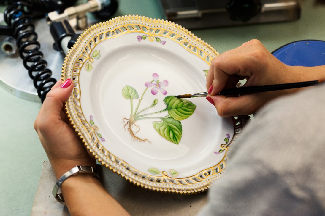 Hands painting a Royal Copenhagen Flora Danica dinner plate with botanical flowers and gold trim.