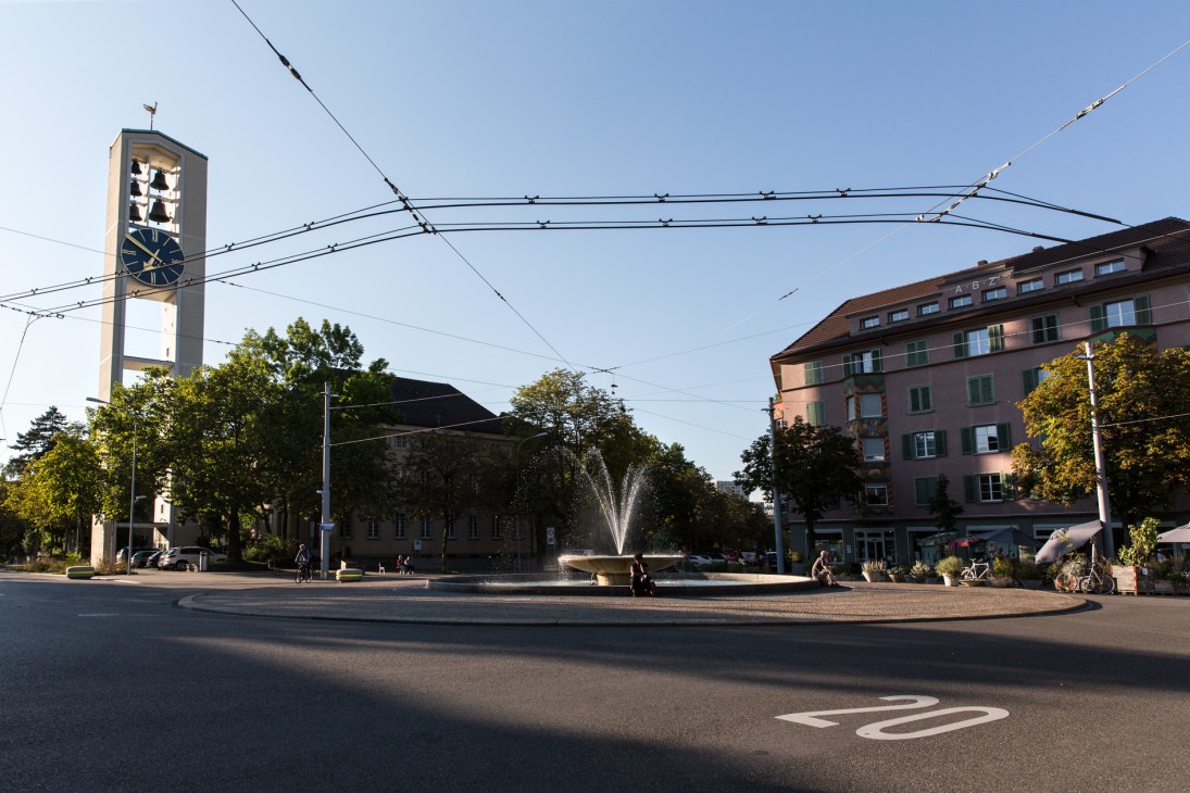 A public square in Zürich's Kreis 4 with a fountain, clock tower, historic buildings, and people enjoying the outdoor space on a sunny day.
