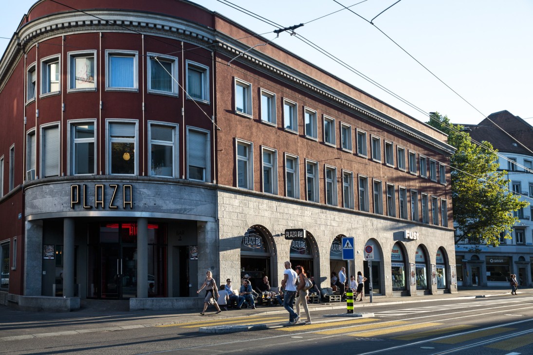 Daytime exterior of nightclub Plaza, a red-brick corner building with large windows and ground-level arches housing retail spaces.