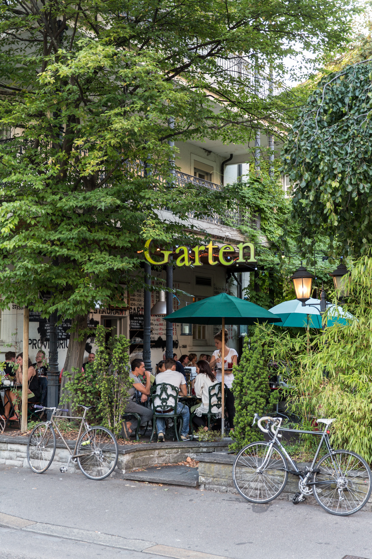 Gartenhof restaurant entrance framed by lush green vines with diners seated under turquoise umbrellas.