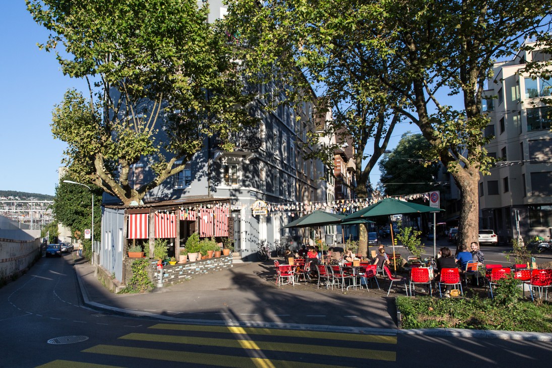 Outdoor café seating with red chairs beneath striped awnings and mature trees on a sunny street in Zürich.