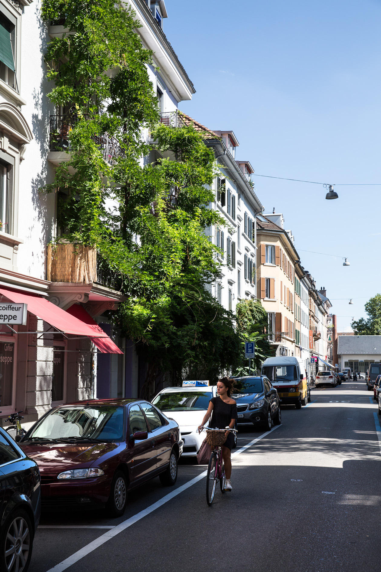 Woman cycling on a sunny street lined with ivy-covered buildings and parked cars in Zürich's Kreis 4 neighbourhood.